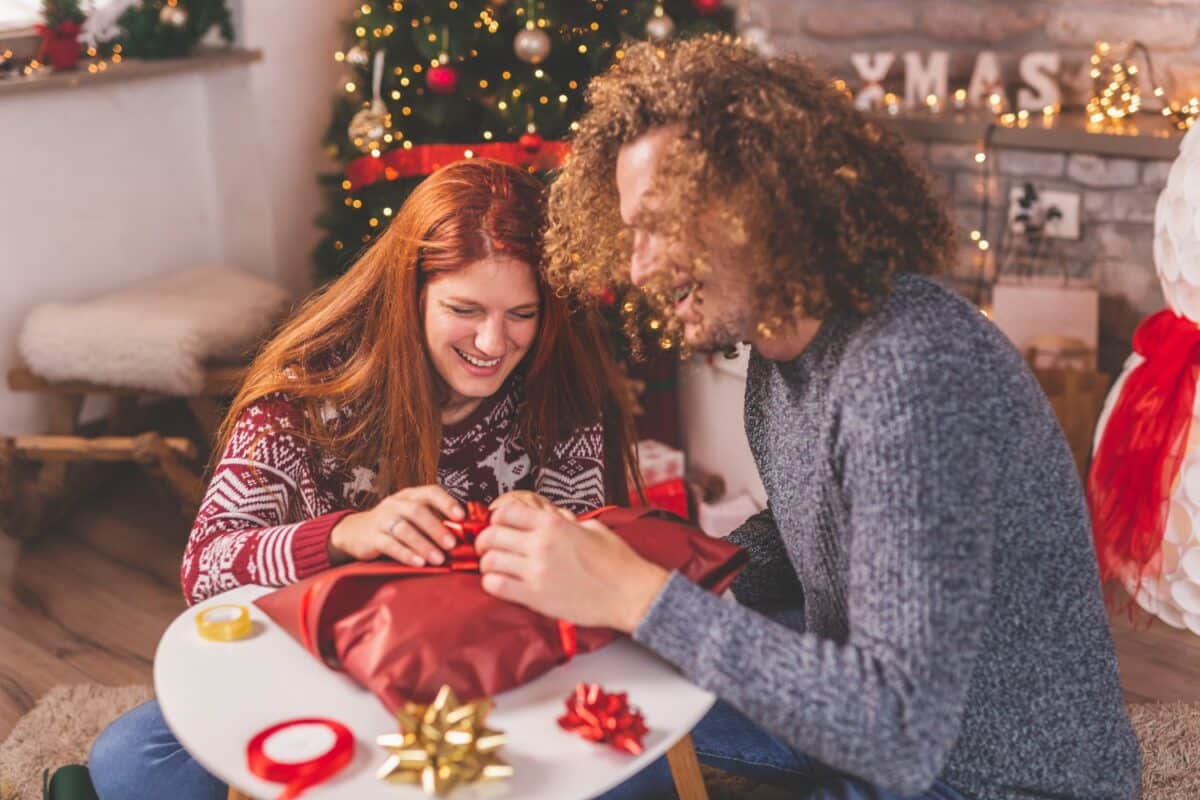 Couple in love sitting by nicely decorated Christmas tree, having fun while wrapping Christmas presents for friends and family