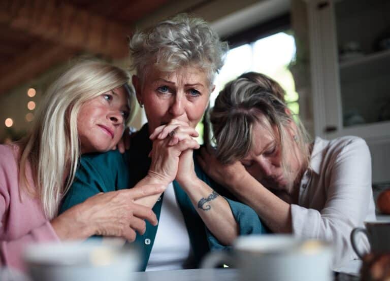 Female senior friends consloing a sad friend indoors at home.