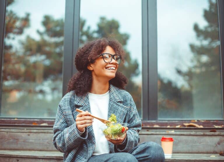 Happy african american woman office worker eating salad and smiling at camera while sitting on bench in park outdoors, selective focus. Positive black girl having lunch outside during break at work