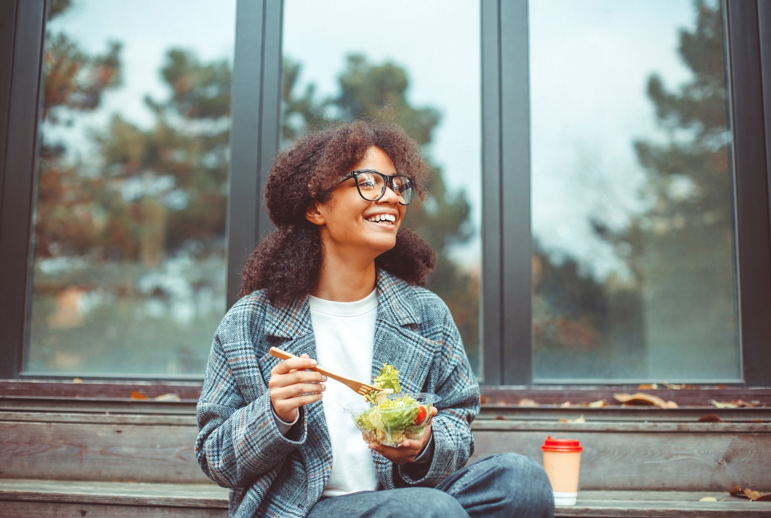 Happy african american woman office worker eating salad and smiling at camera while sitting on bench in park outdoors, selective focus. Positive black girl having lunch outside during break at work