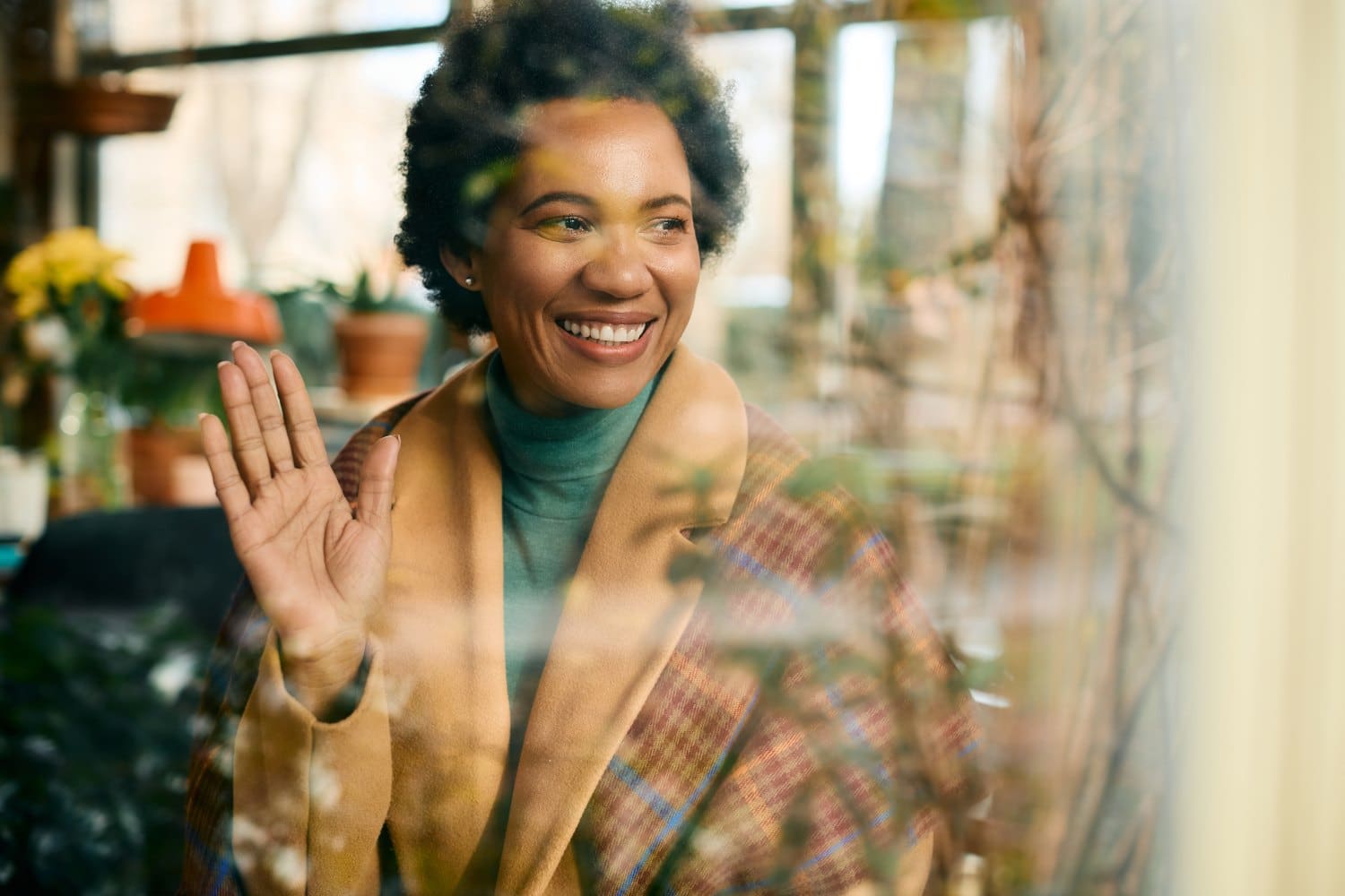 Happy African American woman greeting someone and waving while looking through window of a cafe.