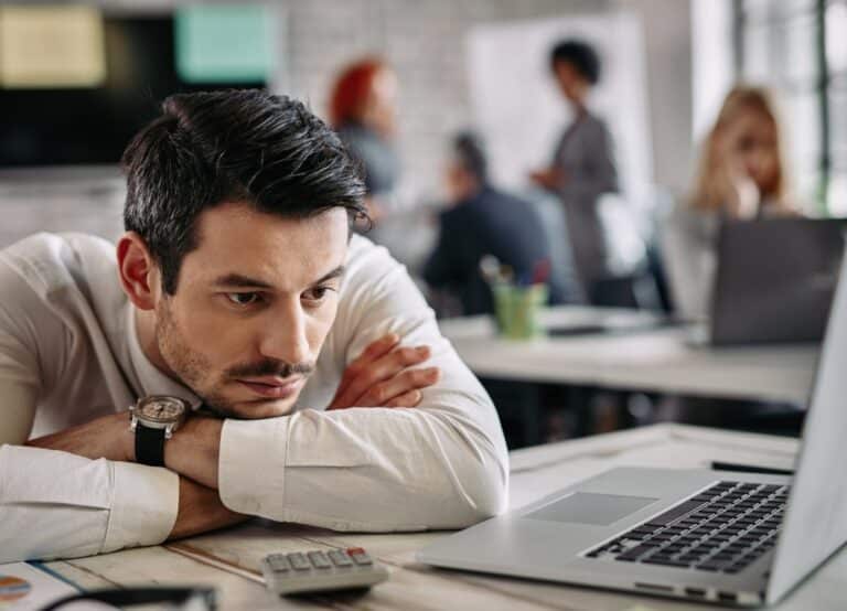Young businessman thinking of something while leaning on his desk and using computer at work. There are people in the background.