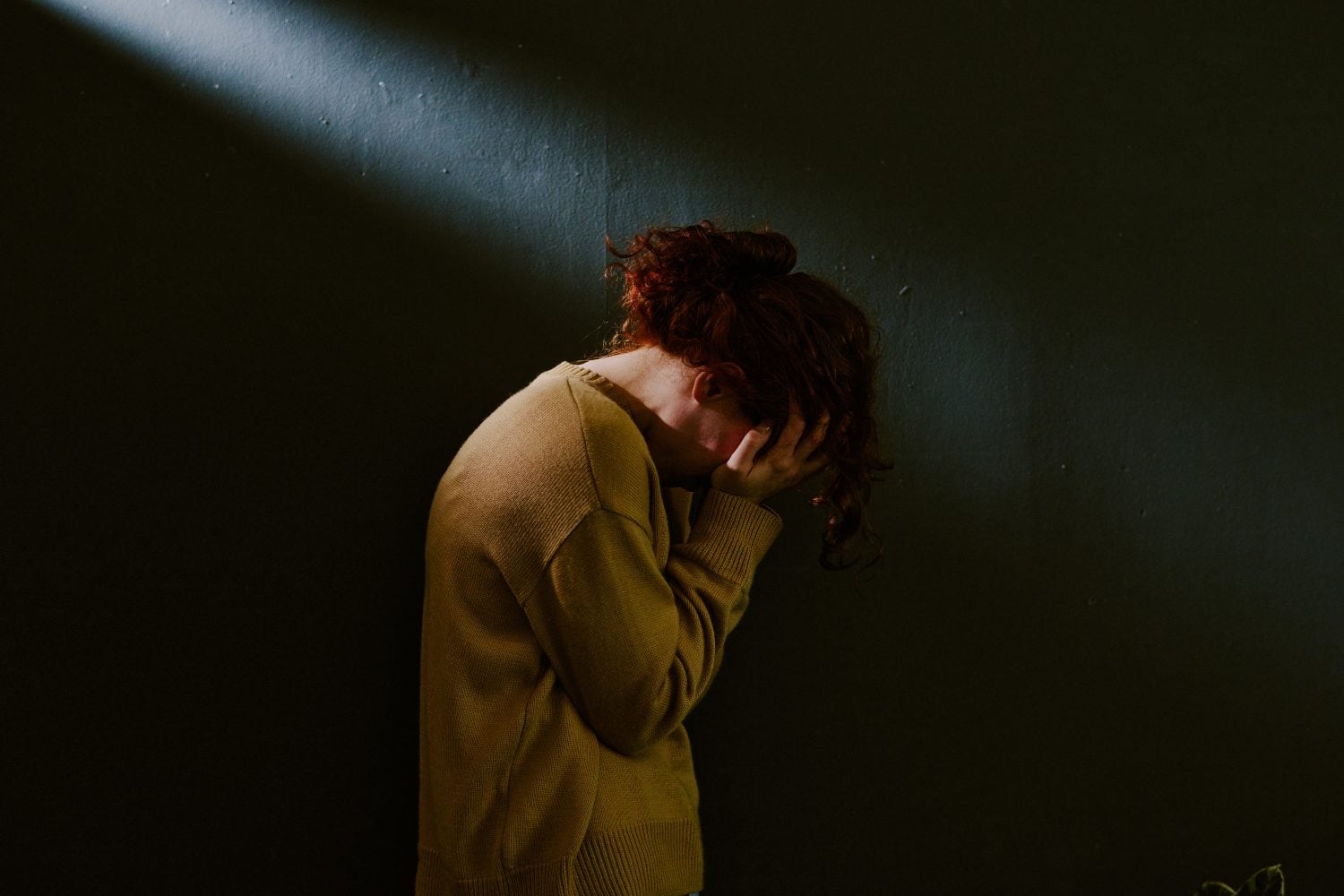young woman with head bowed, wearing sweater, standing near dark wall with light streaming from top left corner, creating dramatic atmosphere filled with solitude