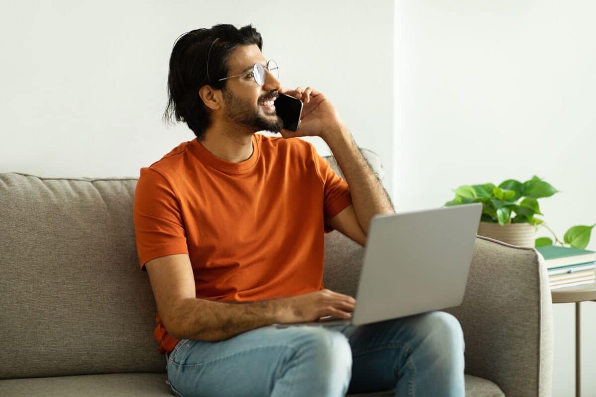 Positive millennial middle eastern man in glasses calls by phone, speaks with laptop, looks at free