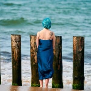 Young blue-haired woman in long dark blue dress with cute tattoo on her shoulder blade standing on sandy beach looking at sea horizon, warm coastal waves caress her feet. Freedom concept (Young blue-haired woman in long dark blue dress with cute tattoo