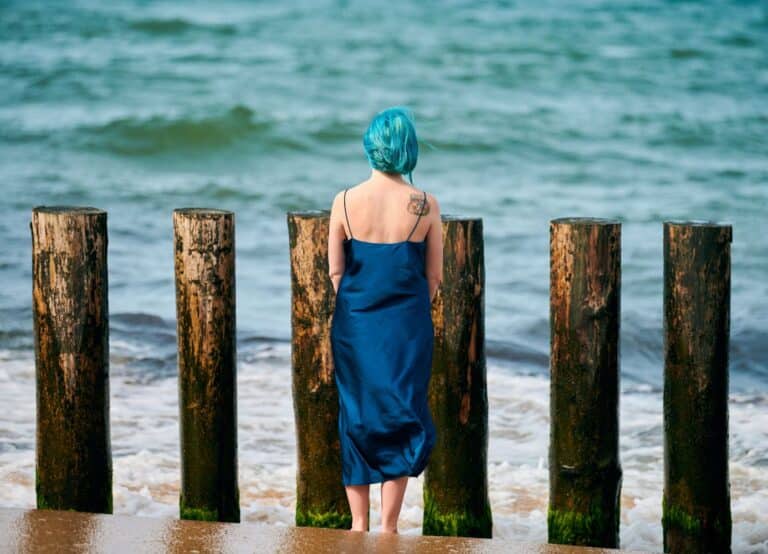 Young blue-haired woman in long dark blue dress with cute tattoo on her shoulder blade standing on sandy beach looking at sea horizon, warm coastal waves caress her feet. Freedom concept (Young blue-haired woman in long dark blue dress with cute tatto