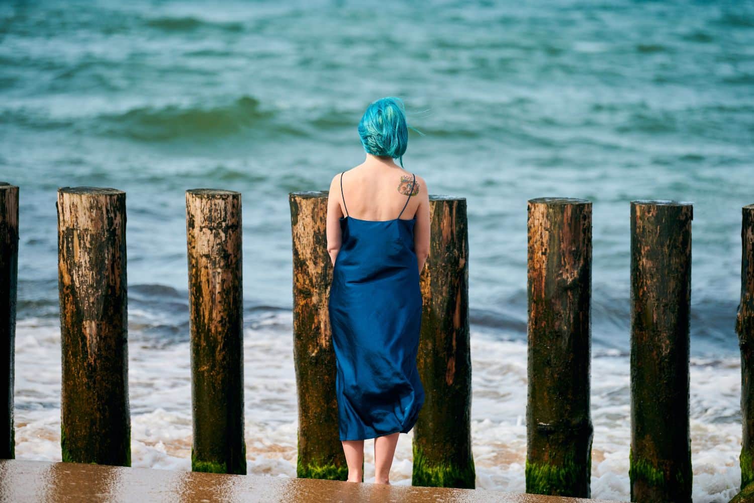 Young blue-haired woman in long dark blue dress with cute tattoo on her shoulder blade standing on sandy beach looking at sea horizon, warm coastal waves caress her feet. Freedom concept (Young blue-haired woman in long dark blue dress with cute tattoo