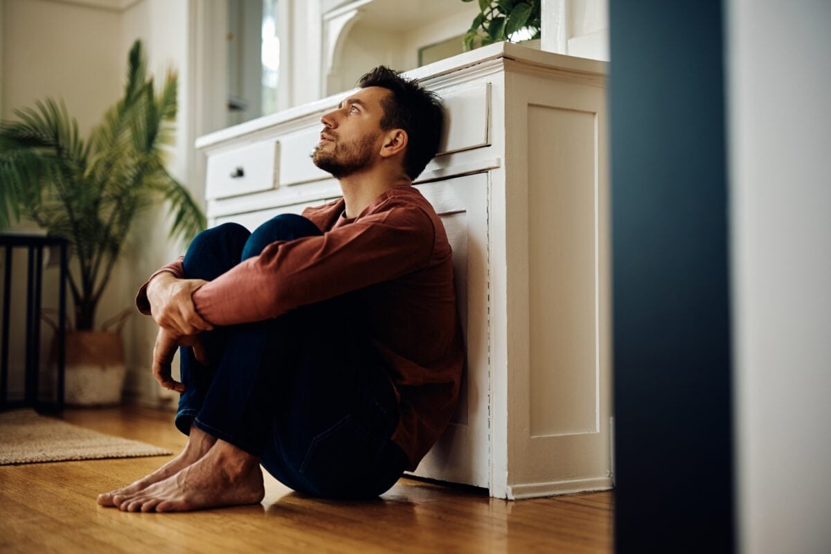 man with dark hair sits on the floor with his knees pulled up to his chest, concept of depression
