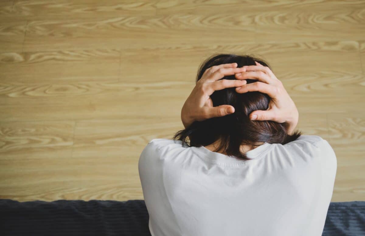 woman sits on a gray sofa in front of a light brown, wood-look wall with her hands on her head