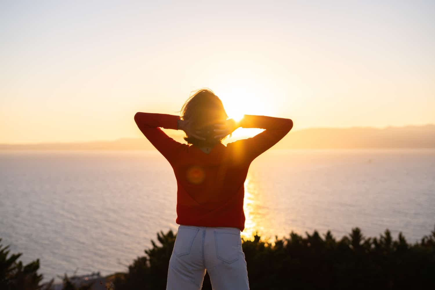 Young woman in a red sweater at a magnificent sunset on the mountain against the background of the sea, romantic mood, back to the camera, freedom